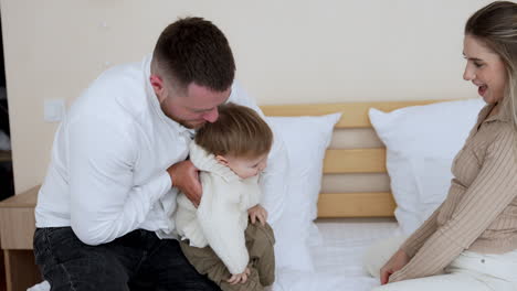 caucasian parents sit on a big bed playing with their toddler son. father holds his little baby boy kissing him with love. happy family portrait.