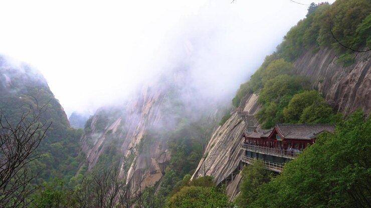A high-angle panning shot of a traditional Chinese-style gallery walkway built precariously along a sheer granite cliff on Mount Hua, surrounded by rolling white fog and green forests.