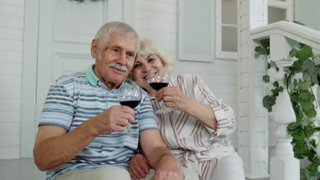 Attractive-senior-elderly-Caucasian-couple-sitting-and-drinking-wine-in-porch-at-home,-making-a-kiss