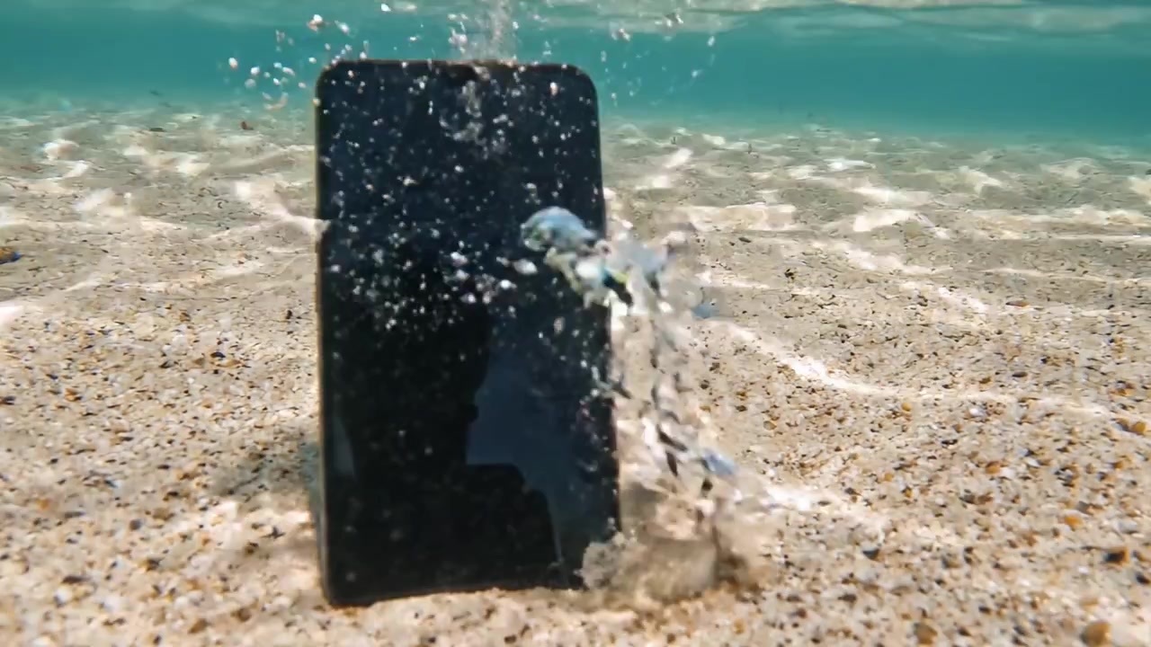 Uno smartphone sommerso giace sul fondale marino, parzialmente coperto da onde leggere e acqua limpida, catturando l'essenza dell'interazione della tecnologia con la natura.