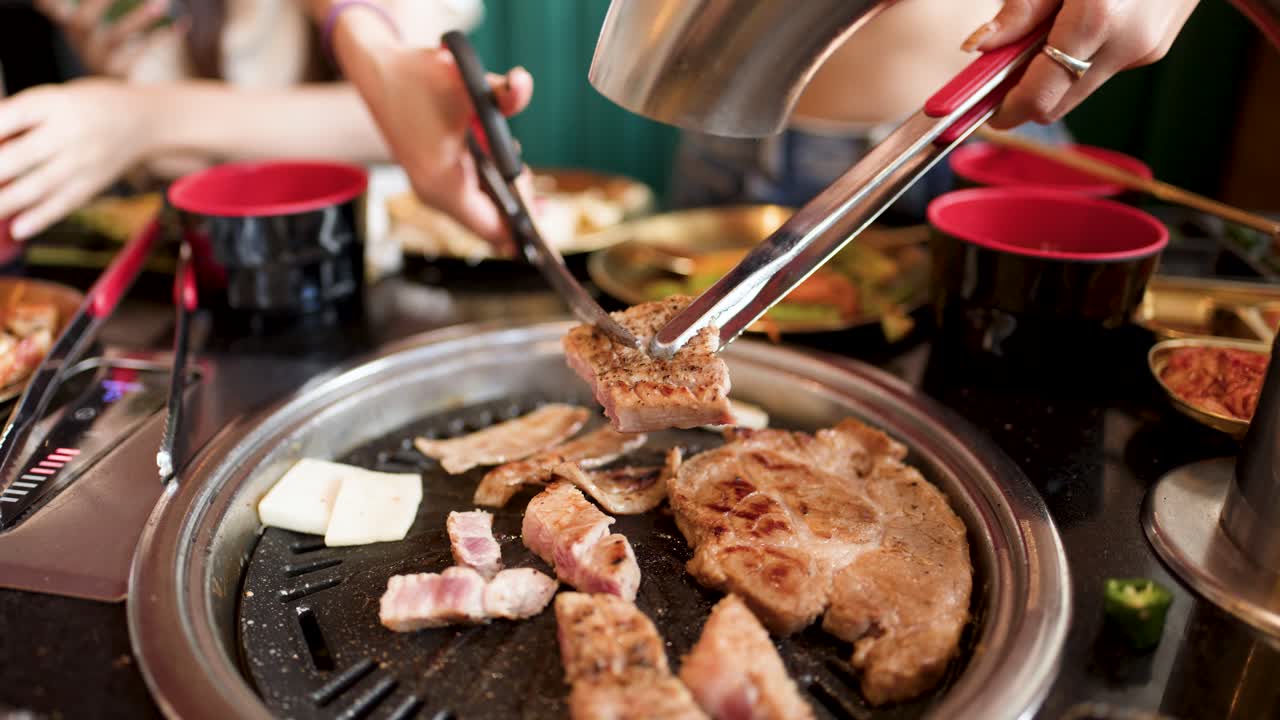 Person slices pork belly with tongs and scissors on tabletop Korean barbecue grill, bright lighting