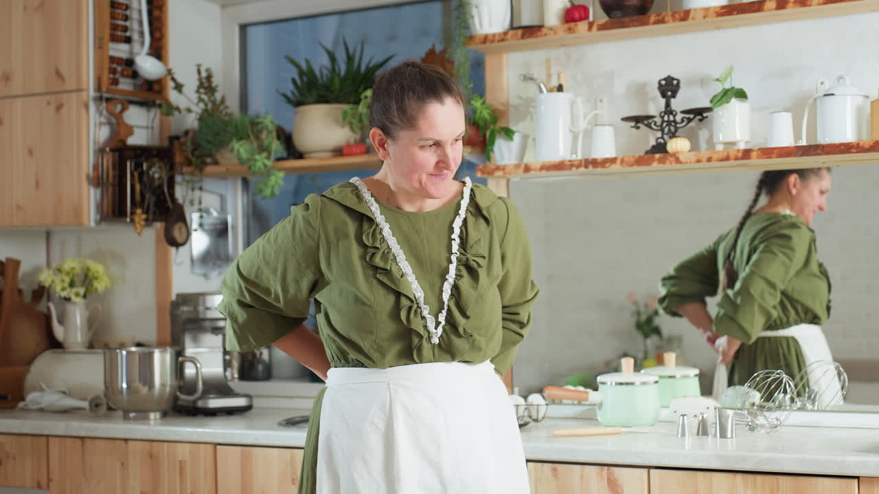 Woman in green dress ties apron around waist in cozy kitchen with wooden cabinets, potted plants, kitchen tools, and mirror reflection visible behind her as she prepares for cooking activity