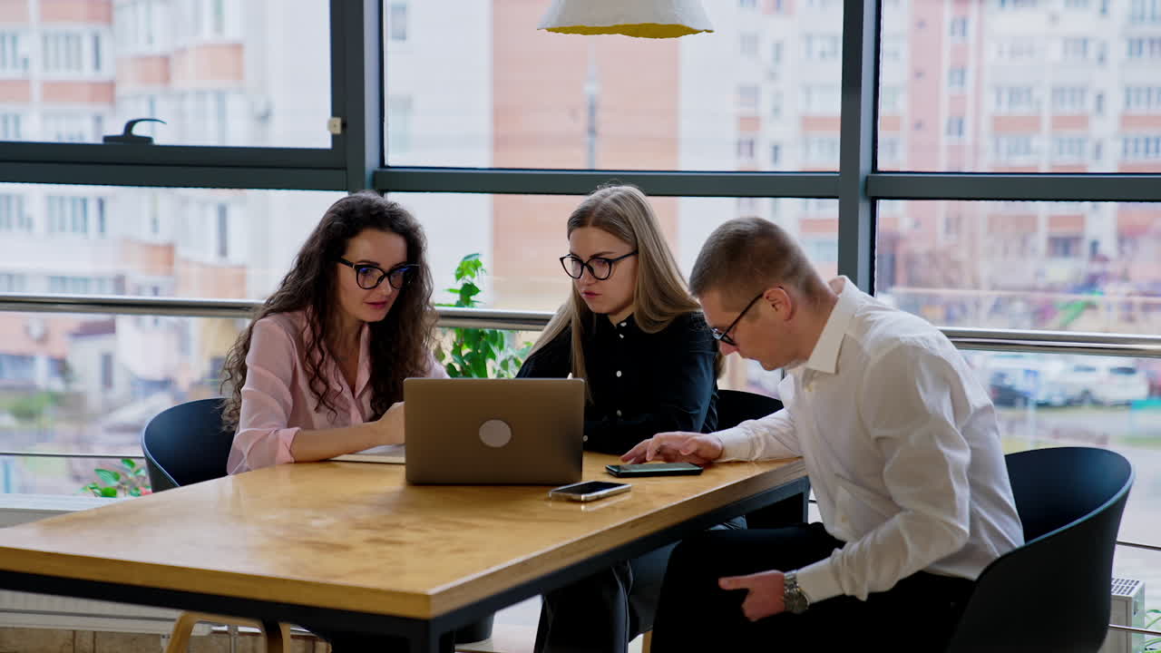 Young coworkers sit at desk look at phones and computer talking. Office employees at work. Windows with cityscape at backdrop.