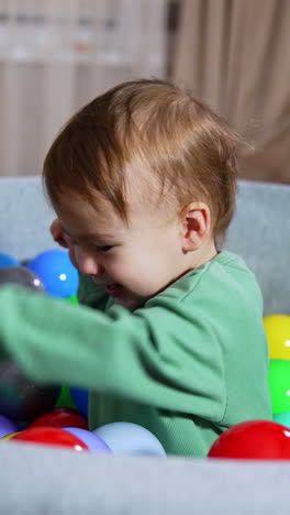 beautiful baby boy sitting in the basin with bright colorful plastic balls. happy child waving hands excitedly. black cat looking wide-eyed and little shocked at backdrop. vertical video