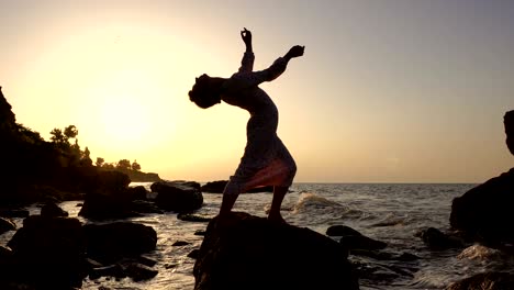 adorable caucasian brunette girl in light dress dancing at the rock in the sea at sunrise time