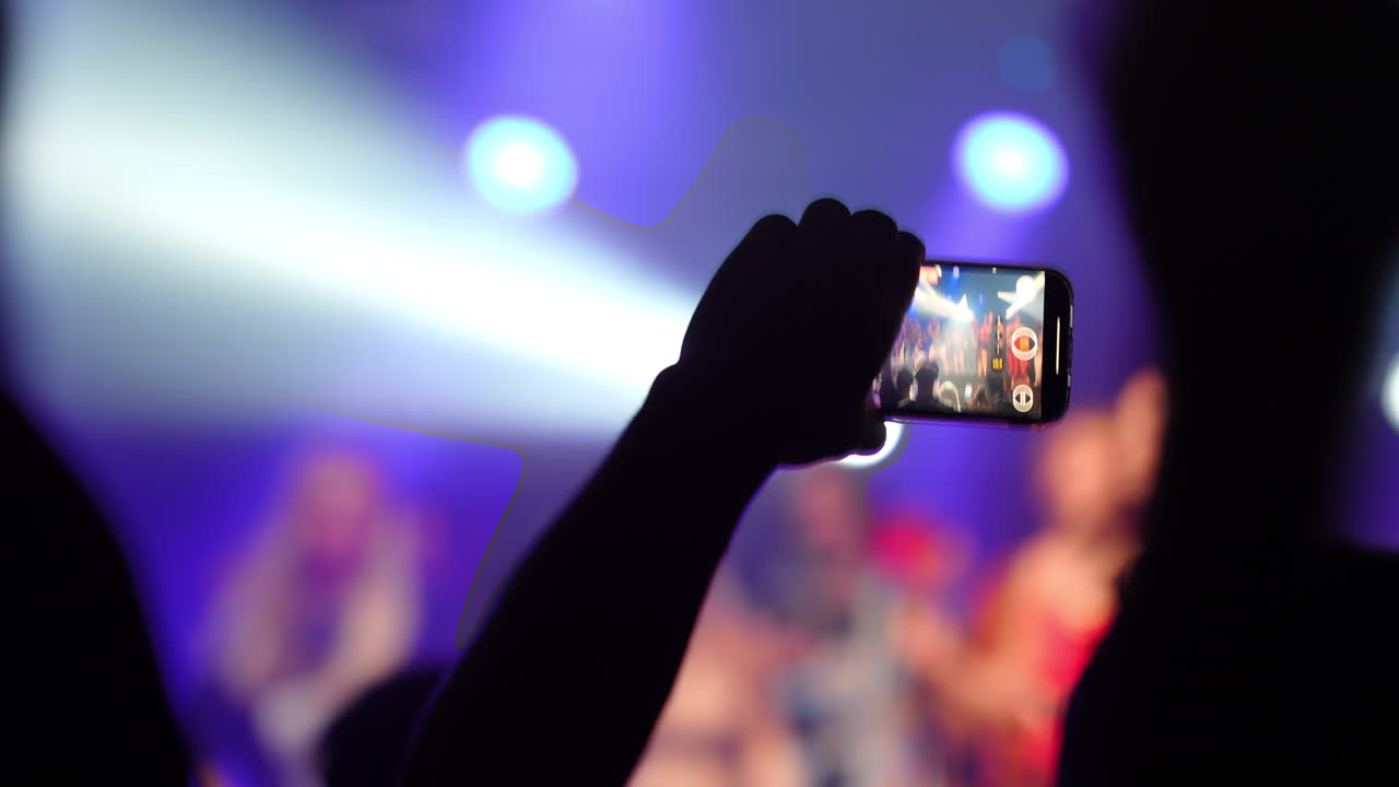 Spectator capturing a live music concert with a smartphone camera, surrounded by vibrant stage lights and an enthusiastic crowd, creating an electrifying atmosphere filled with energy