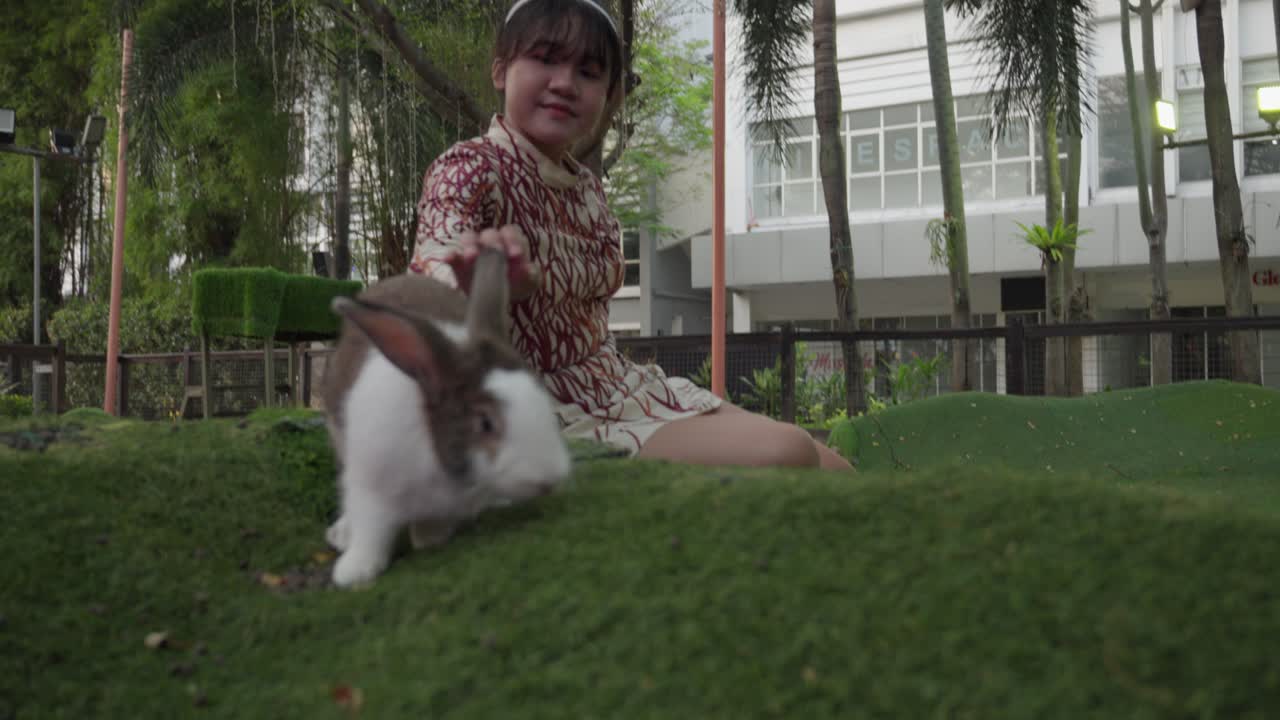 A happy young girl sits on the grass while playing with her pet rabbit in a park setting
