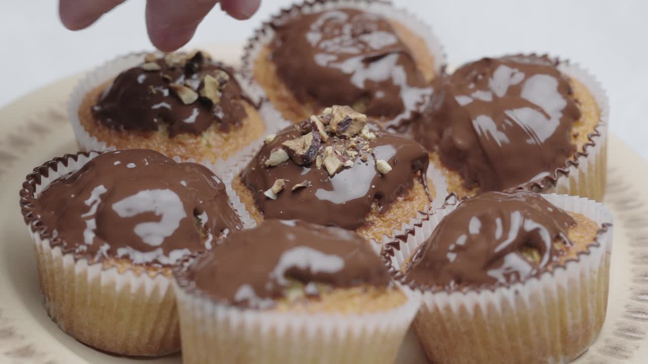 Una mano aggiunge noci tritate su muffin glassati al cioccolato in un primo piano della preparazione di un dolce.