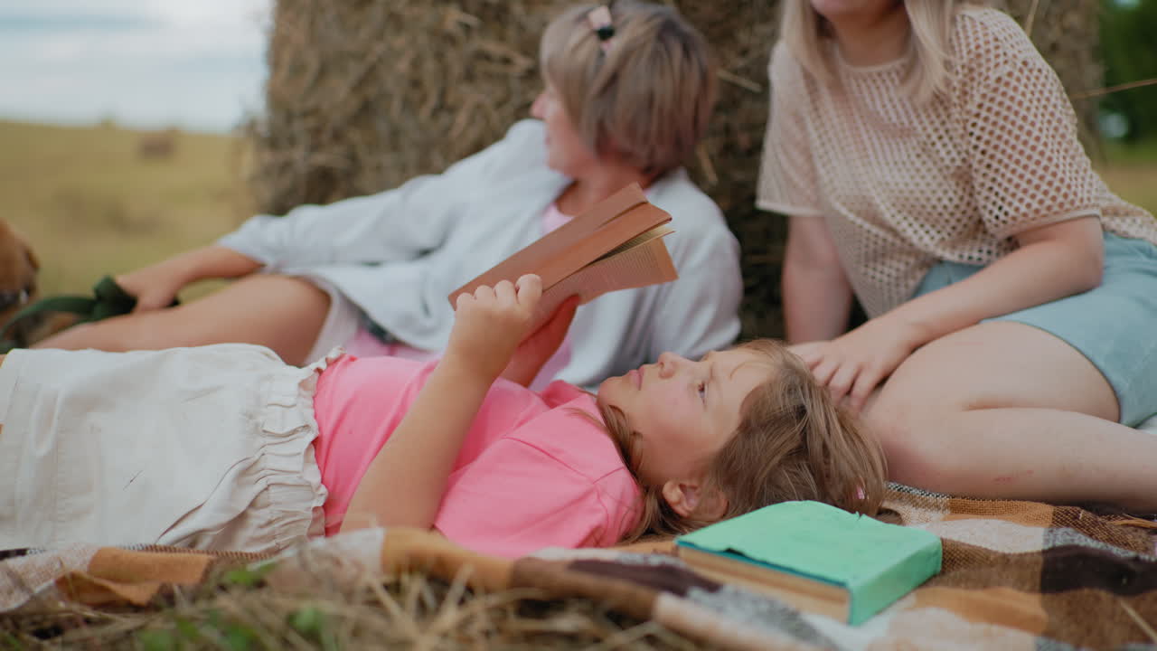 핑크색 셔츠를 입은 어린 소녀가 피크닉 담요 위에 누워서 초록색 표지판으로 책을 읽는 클로즈업, 어머니와 할머니가 가까이 앉아, 개가 에 휴식을 취하고, 시골에서 야외에서 가족 시간을 즐기고 있습니다.