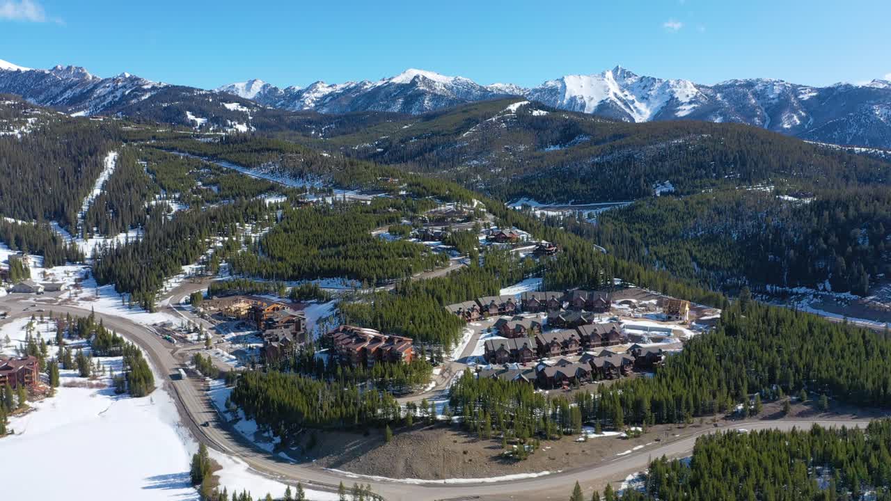 Aerial drone footage of Big Sky, Montana in winter shows cozy condos nestled among pine trees with snowy rooftops and breathtaking mountain views surrounding this peaceful alpine town