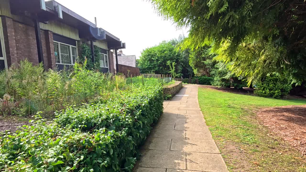 イギリスのダラムにある、豊かな緑と手入れの行き届いた景観に囲まれたレンガ造りの建物の脇を、日当たりの良い庭の小道に沿って安定したカメラが移動する。