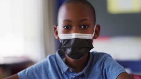 portrait of african american schoolboy wearing face mask, sitting in classroom looking at camera