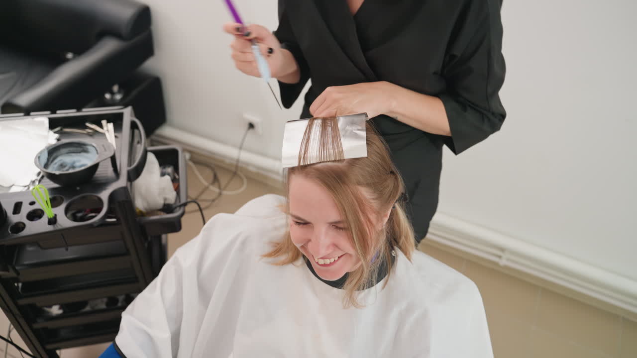 Salon Highlights Folien Anwendung lächelnde Frau, Coloristin trägt mit Pinsel und Kamm Aluminiumfolien und Farbe auf mittellanges blondes Haar auf, Kundin sitzt in weißem Umhang, Salonwagen mit Schalen
