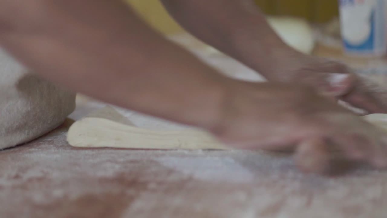 Close-up of a pastry chef rolling dough with hands using a wooden rolling pin on a work surface.