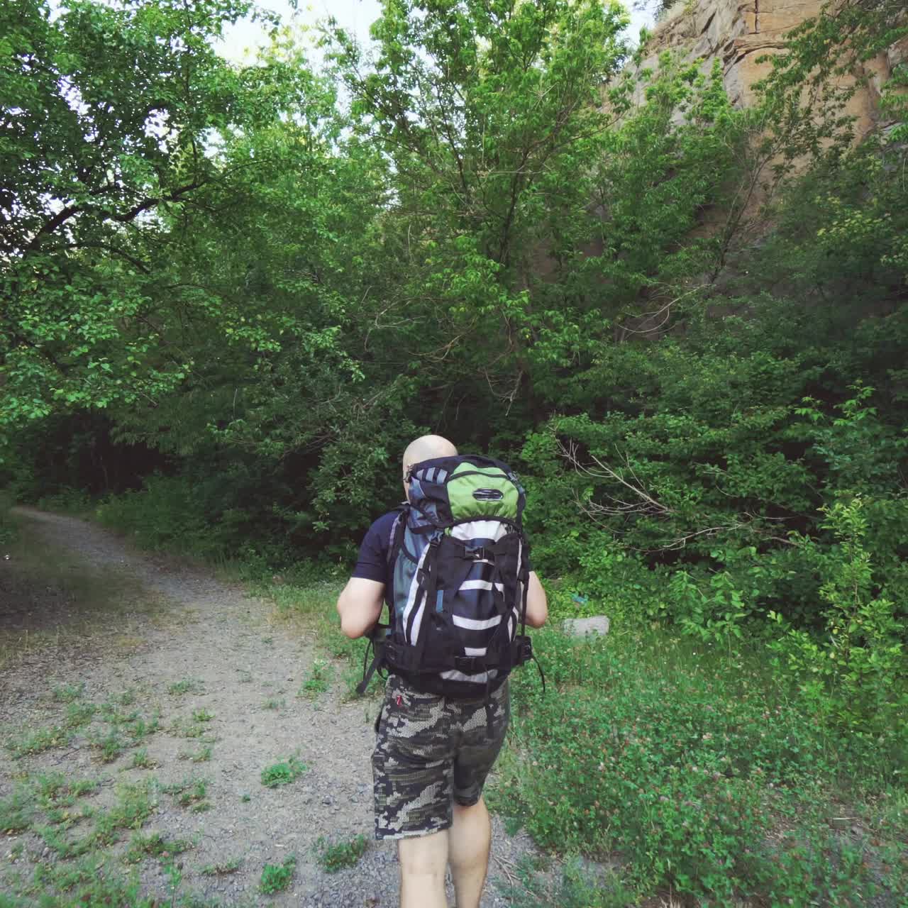 tourist with a backpack in shorts and a t-shirt is going along a path in the forest near the rock not far from precipice in a warm summer day