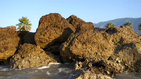 Nahaufnahme-Der-Schäumenden-Wellen-Der-Adria,-Die-Gegen-Die-Felsige-Küste-Eines-Albanischen-Strandes-Krachen,-Mit-Palmen-Im-Hintergrund
