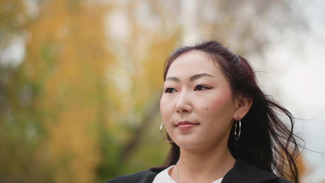 長い黒髪の若い女性のクローズアップ。彼女は大きなフープピアスと白いブラウスの上に黒いブレザーを着用し、穏やかな表情でぼやけた秋の景色に囲まれた屋外に立っている。