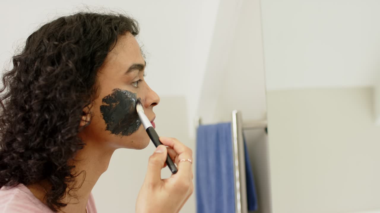 Dipping brush into charcoal mask jar, non-binary person applying across cheek at sink, copy space