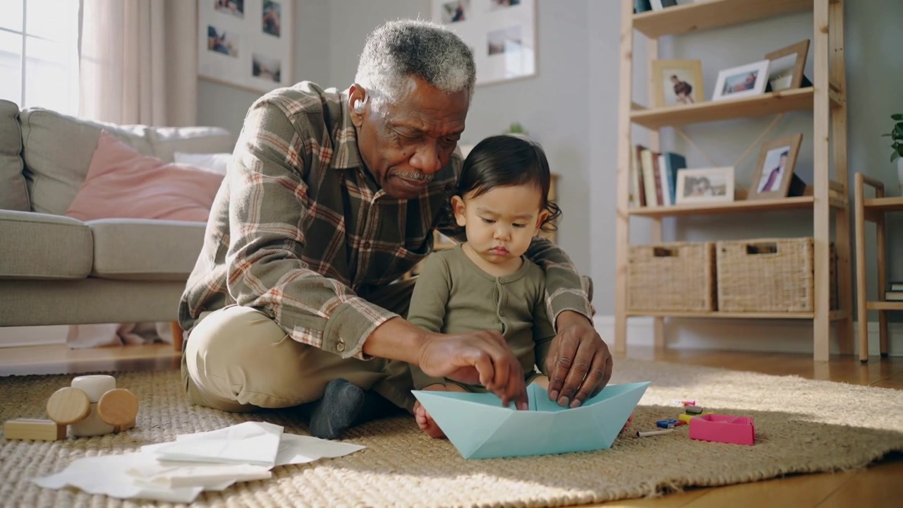 Placing folded blue paper plaid-shirted man teaching olive-onesie toddler folding boat at home