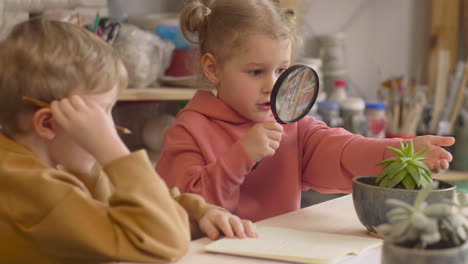 little girl talking with a friends while observing a plant with a magnifying glass sitting at table in a craft workshop