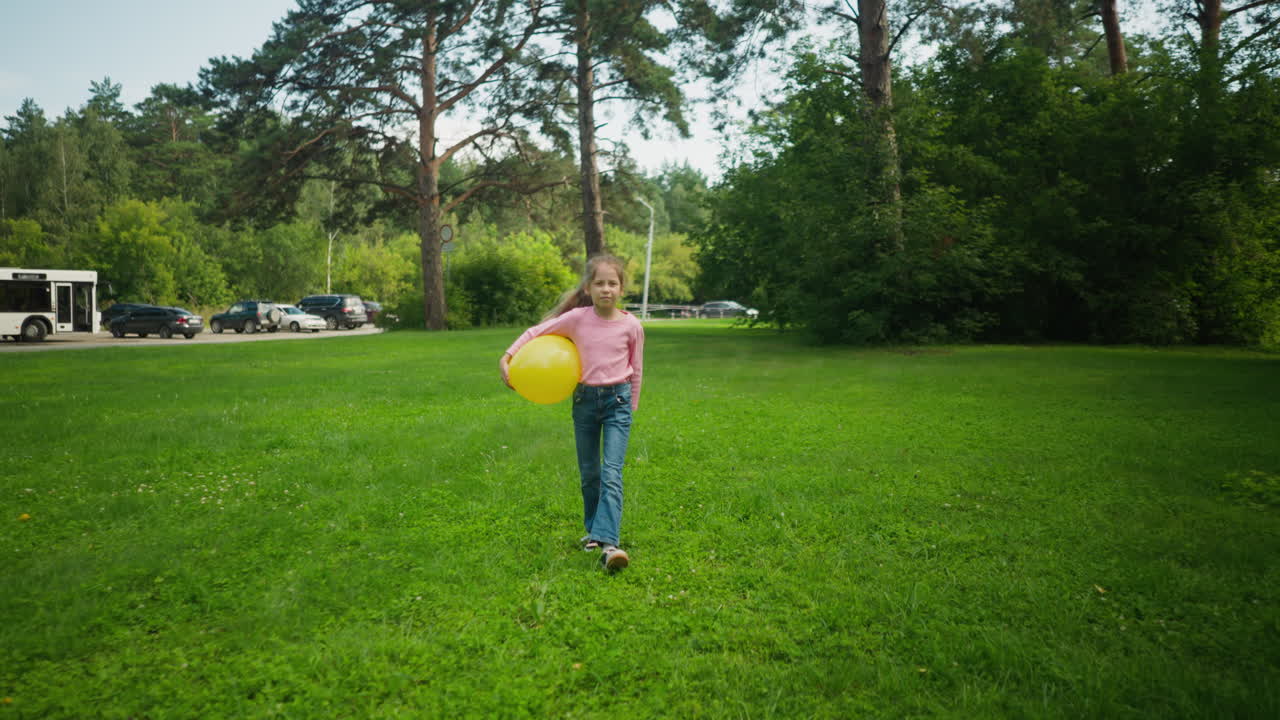Front view of young girl in pink top and blue jeans walking confidently on grassy field holding bright yellow balloon under arm, with trees and parked cars in background under clear daylight