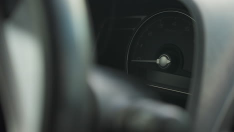 close up of car tachometer showing idle engine rpm with ignition off, blurred steering wheel partially frames instrument cluster, odometer and warning light visible inside dimly lit dashboard panel