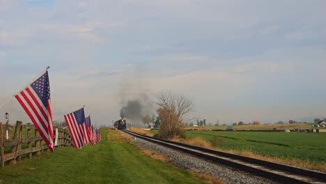 una vista de una sola vía ferroviaria, con una valla con la bandera de américa en ella, agitando suavemente en el viento a medida que se acerca un tren de vapor, en un soleado día de otoño