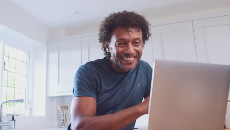 Mature-Man-At-Home-In-Kitchen-Waving-As-He-Makes-Video-Call-On-Laptop-Computer