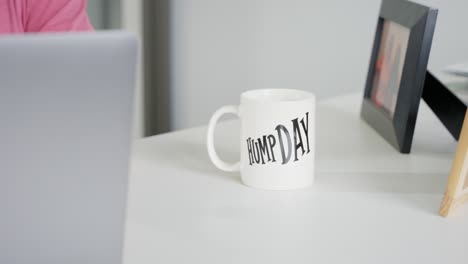 glass mug with hump day print on white office table, workplace with employee on laptop