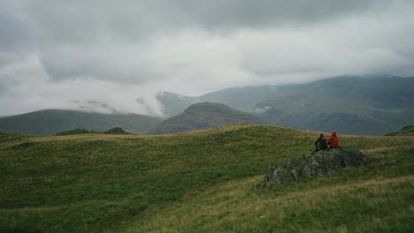 two hikers in jackets embrace on a rock watching clouds blow over the mountains