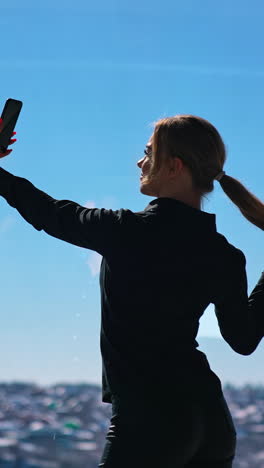 lovely business woman in black taking photo of herself. beautiful young woman doing selfie on smartphone while standing at office window. vertical video