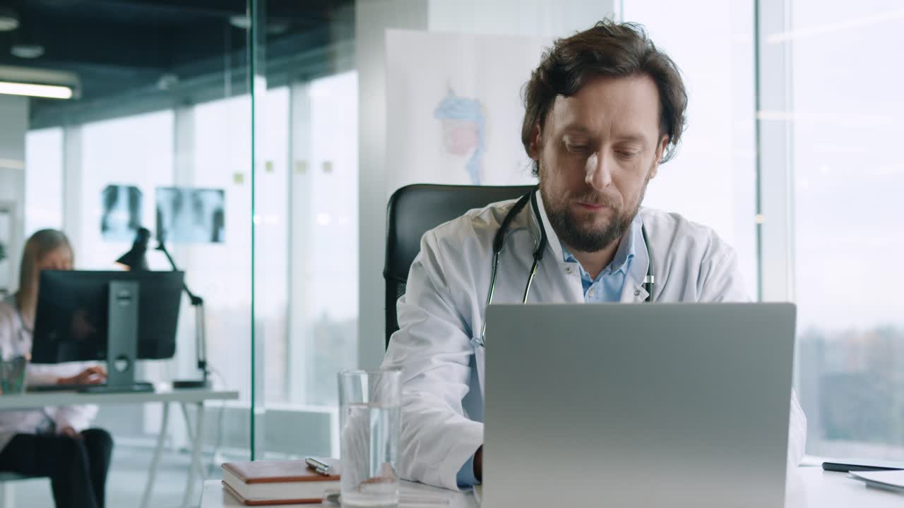 vista de primer plano del médico varón caucásico senior sentado en la oficina del hospital trabajando y escribiendo en la computadora portátil en el lugar de trabajo