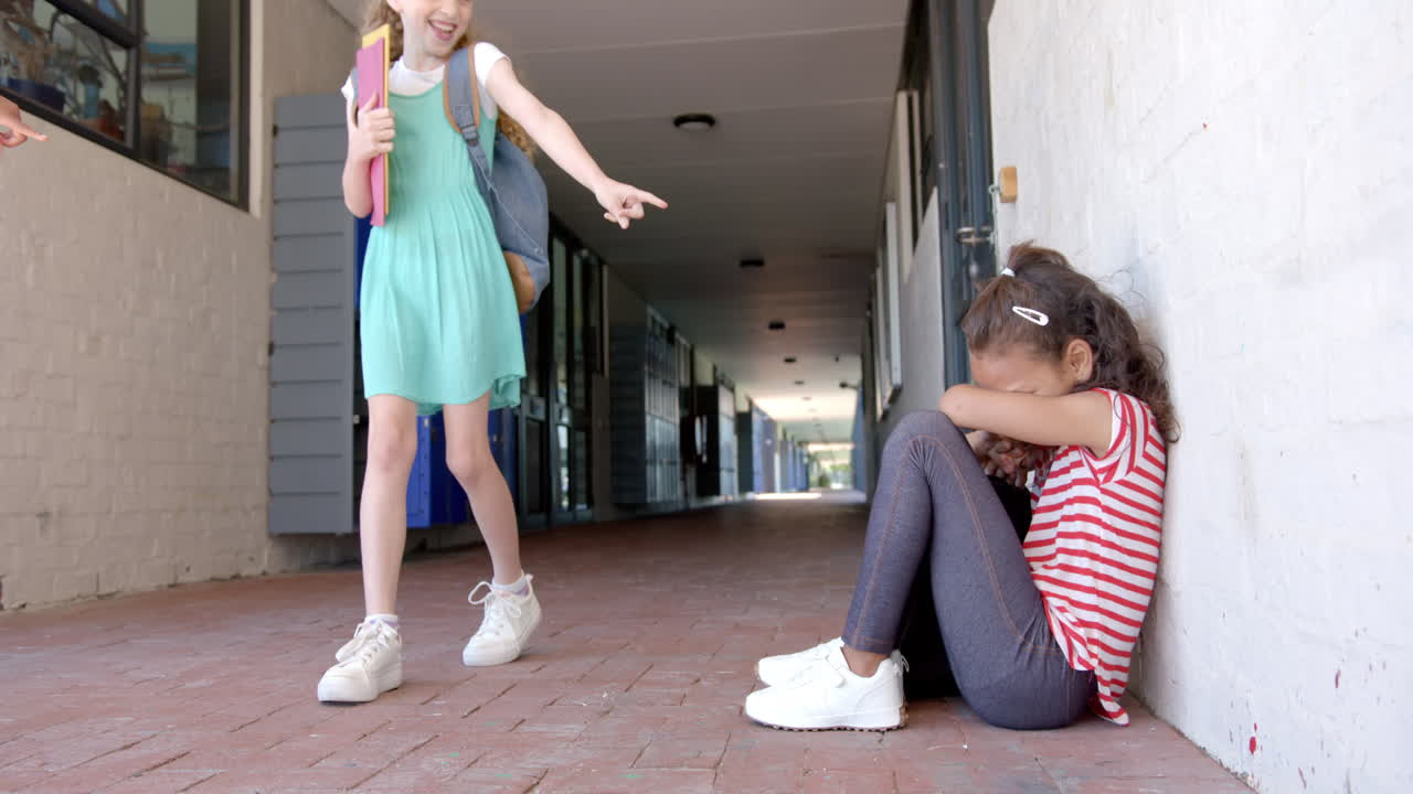 In school, girl pointing at another girl sitting on floor, feeling upset