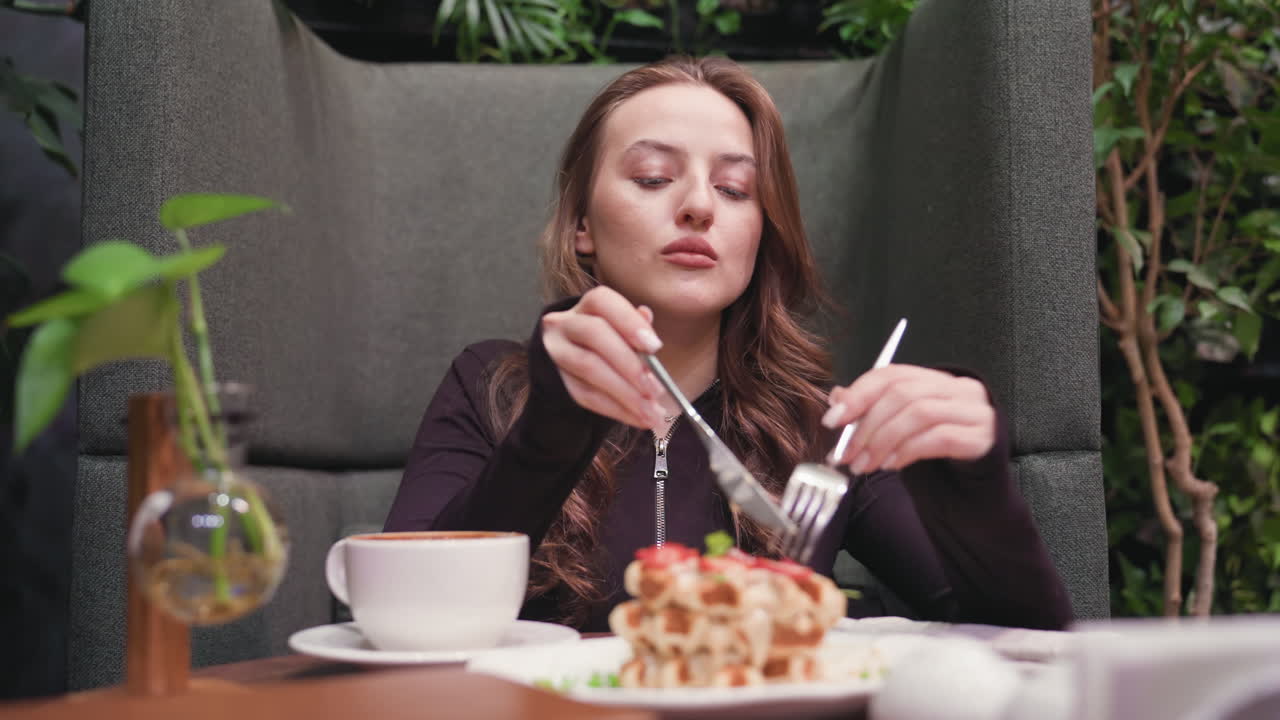 Lady seated in cozy cafeteria booth preparing to cut delicious stack of waffles with knife and fork, surrounded by greenery, glass vase with plant, hot latte in white cup, and stylish table setting