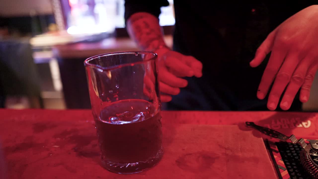 Tattooed bartender stirs cocktail in glass, dim bar setting, warm red ambient lighting, close-up
