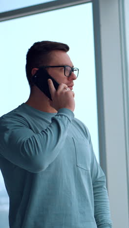 caucasian fit man standing at the panoramic window with hand in the pocket. businessman having phone conversation looking at the window. vertical video
