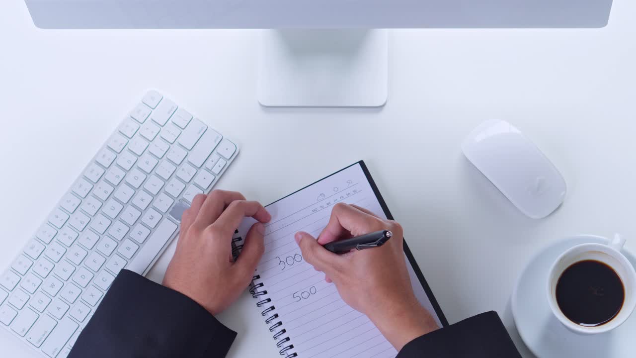 Top view of businessman writing notes in spiral notebook at clean white desk with computer