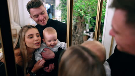 beautiful caucasian couple stand holding their infant in front of the mirror. mom plays with her son and kid smiles adorably.