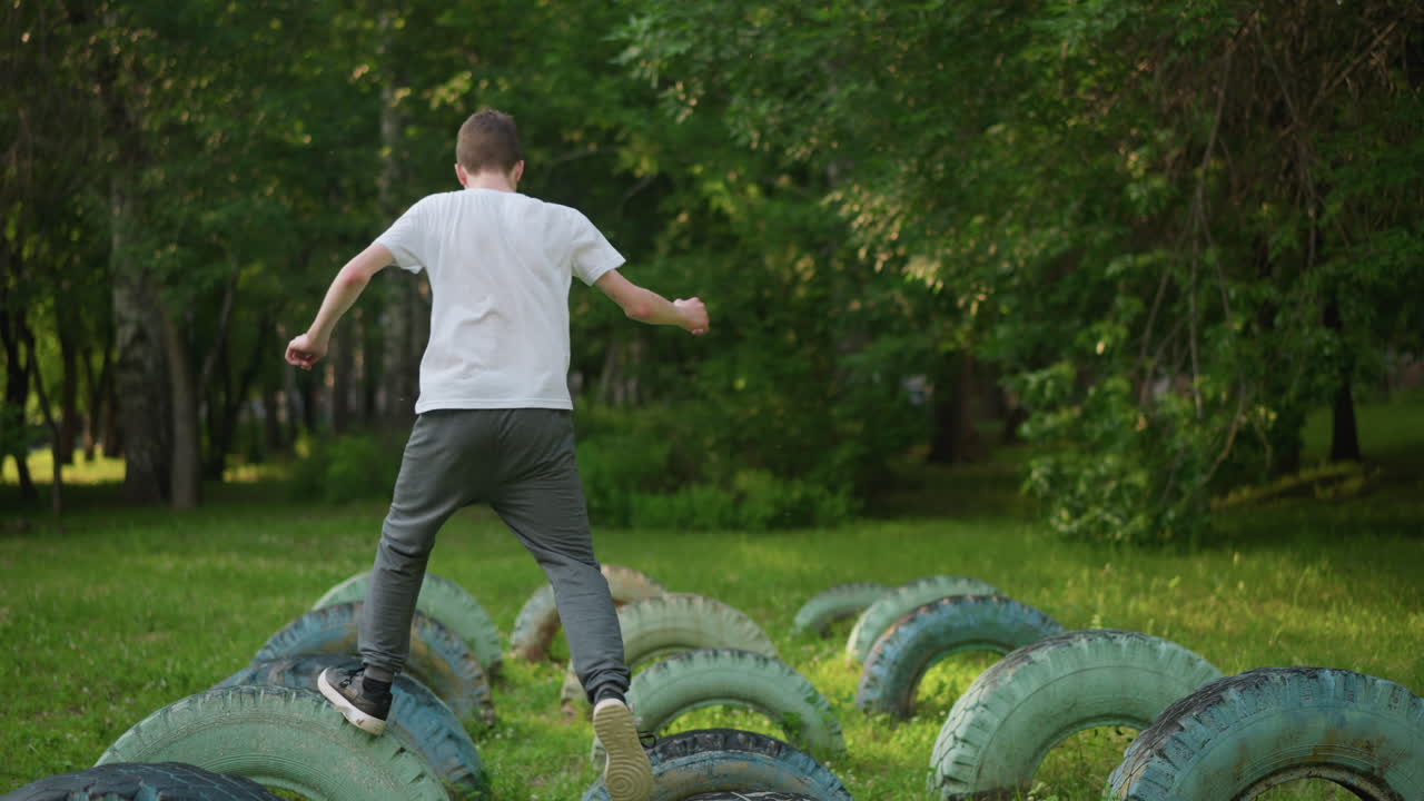 Slow-motion back view of a young boy in a white shirt and gray pants training on a series of tires in a grassy field, surrounded by trees in the background