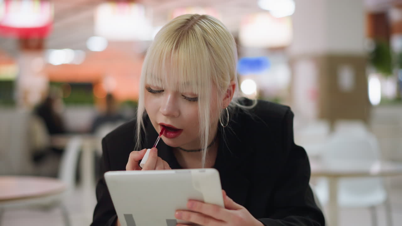 Beauty model applying lipstick while holding tablet as mirror in indoor cafe, focusing on lips with calm expression, showing makeup routine and personal care with blurred colorful background lights