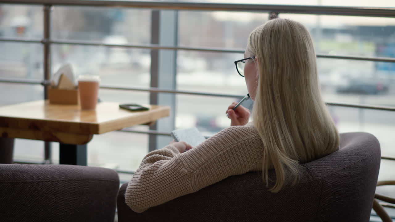 vista trasera de una mujer sentada en una mesa con un libro y una taza de café, escribiendo pensativamente en un cuaderno, mostrando una atmósfera acogedora y enfocada, con un fondo suave de una cafetería o espacio de oficina