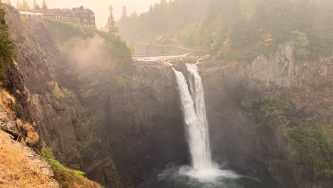 large waterfall in the mountains, next to a cozy, cliffside, lodge on a smoky, cloudy day
