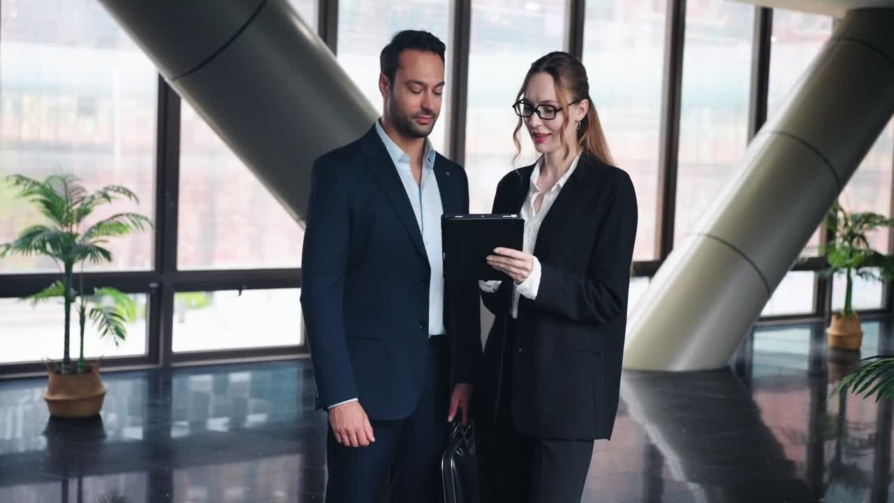 Professional business secretary shows project documents on a digital tablet screen to her boss while standing in a spacious modern corporate office lobby near large windows