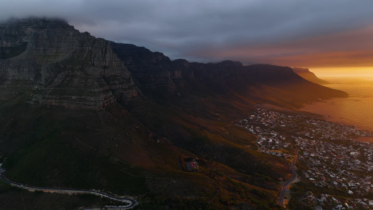Veduta aerea cinematografica della Table Mountain a Città del Capo, Sudafrica, scogliere rocciose drammatiche con paesaggio urbano costiero al tramonto, nuvole che si muovono sulle cime e luce dorata della sera.