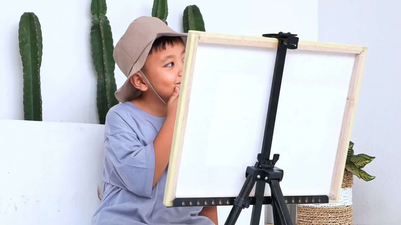 Anak laki-laki muda bertopi dan memakai headphone sedang melukis di kuda-kuda, sangat berkonsentrasi pada karyanya