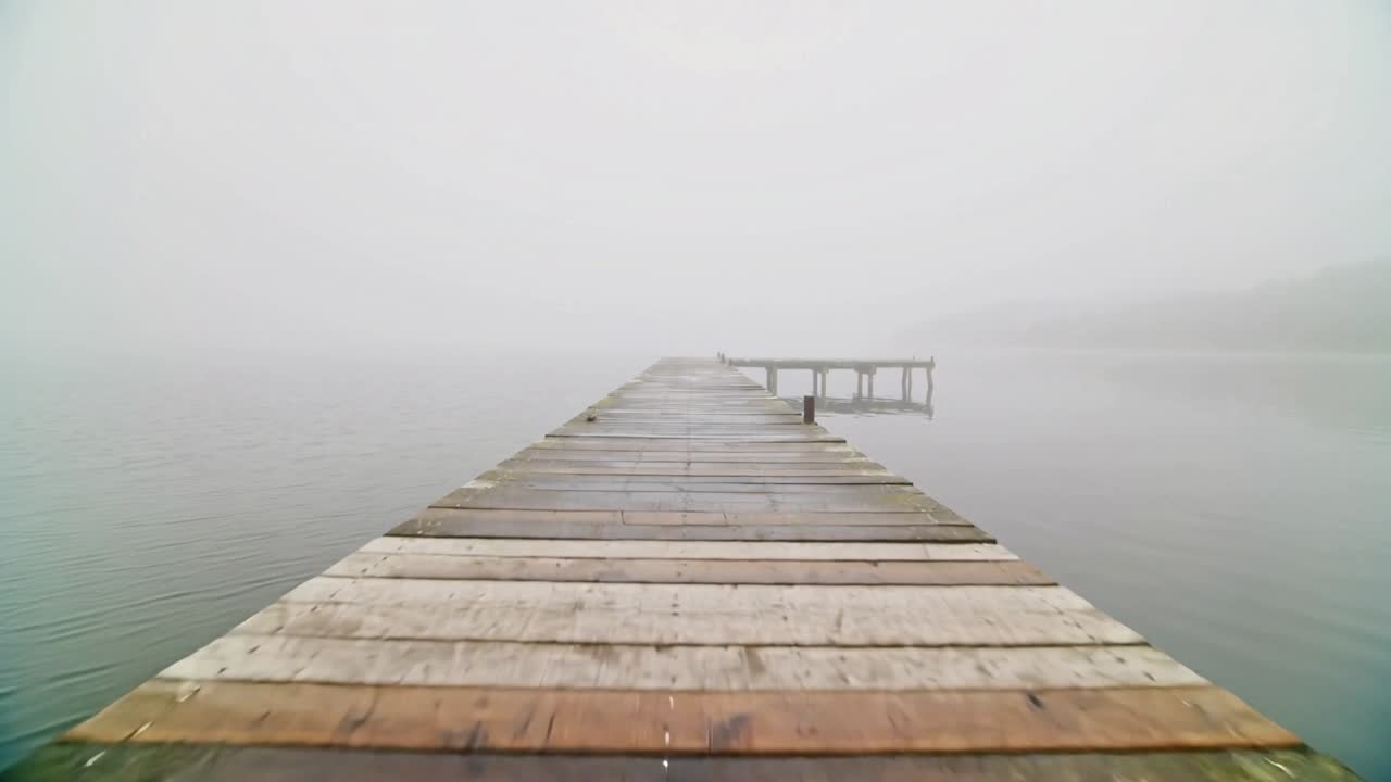 Telecamera in movimento che avanza lungo un pontile di legno usurato su un lago calmo, rivelando una piattaforma distante.