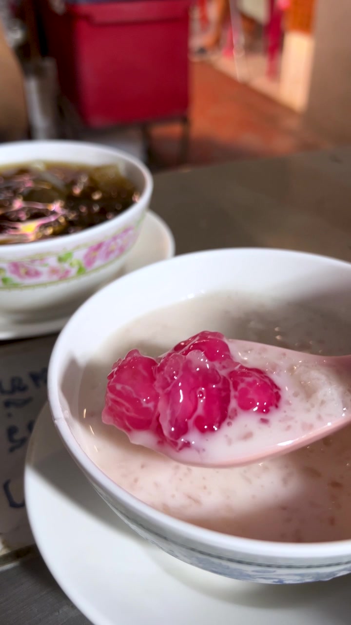Tangan mengangkat sesendok hidangan penutup delima merah dari mangkuk santan di bawah pencahayaan jalan yang terang
