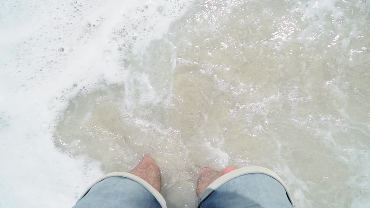 Caucasian Man in Shorts Looking down to his Feet and gets Hit from Wave on Beach