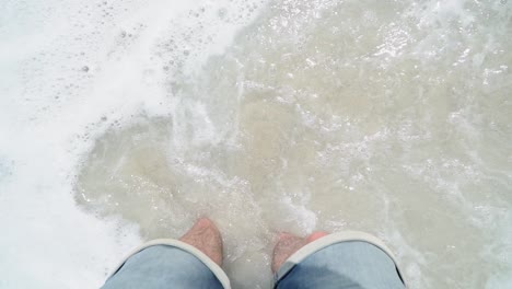caucasian man in shorts looking down to his feet and gets hit from wave on beach