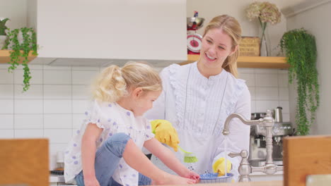 Mother-wearing-rubber-gloves-at-home-in-kitchen-with-young-daughter-having-fun-as-they-do-washing-up-at-sink--shot-in-slow-motion